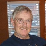 A man with a blue shirt, gray hair, a mustache, and glasses is smiling.